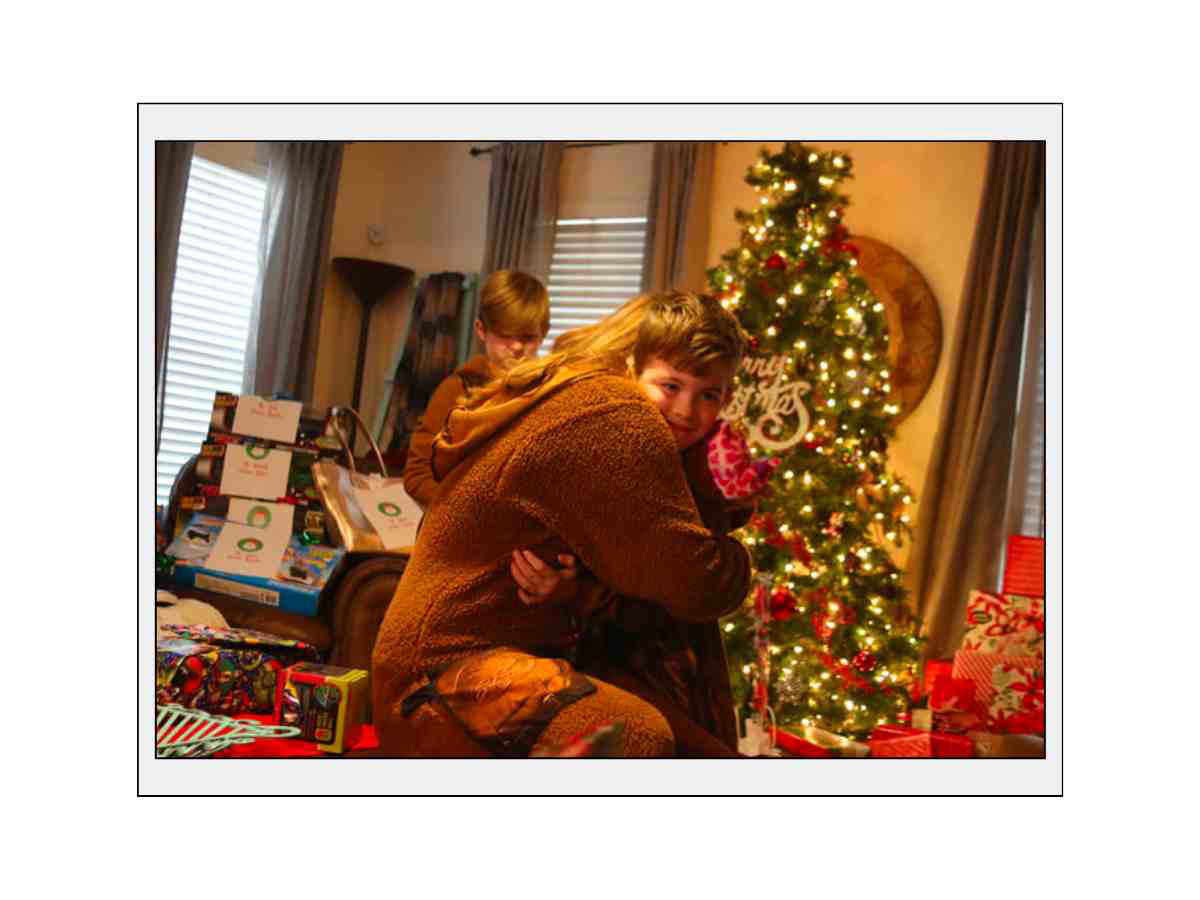 christmas photos to take of siblings Two children hugging in a room decorated for Christmas, with a Christmas tree adorned with lights and ornaments in the background, and several wrapped gifts surrounding them.