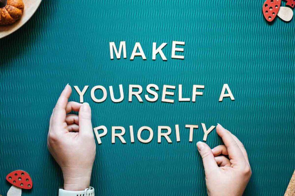 make yourself a priority self care quote Wooden letters on a green textured background spell out 'MAKE YOURSELF A PRIORITY,' with hands arranging the letters.