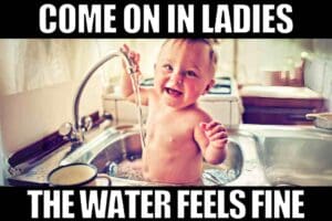 Cheeky baby grinning while sitting in a kitchen sink bath, inviting the ladies in with a splash, embodying the charm of a classic baby meme.