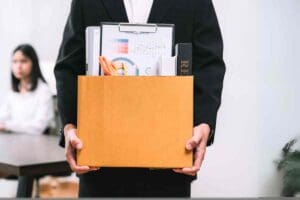 Meme of a person holding a cardboard box filled with office supplies, symbolizing the classic 'last day of work' exit strategy, while a coworker looks on in mild disbelief.