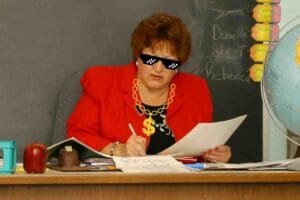 Meme of a teacher in a red blazer wearing pixelated sunglasses and a gold dollarsign chain, looking at papers with a globe and apple on the desk, embodying that gangster educator vibe.