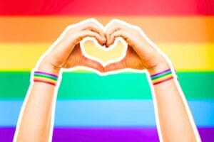Meme featuring hands forming a heart, adorned with rainbow wristbands against a vibrant pride flag background, celebrating love with a side of glitter and giggles for Pride Month.