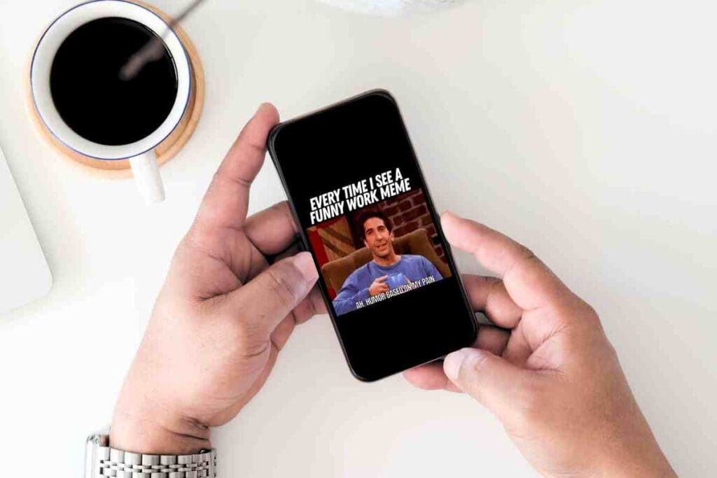 A humorous work meme on a smartphone screen shows a person smirking with text: 'Every time I see a funny work meme... ah, humor based on my pain.' Next to a cup of coffee—perfect for a tired employee seeking a giggle.