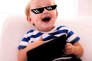 A meme featuring a joyful toddler wearing pixelated sunglasses, holding a tablet and sporting a mischievous grin like he's just become the CEO of adorable antics.