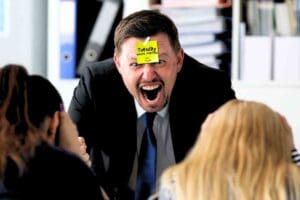 Grinning man in a suit with a 'Tuesday Work Memes' sticky note on his forehead, humorously expressing that Tuesday is as thrilling as a surprise meeting.