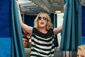 Meme of a woman in sunglasses dramatically opening airplane curtains, embodying all of us saying, 'I am ready for my inflight announcement, but first, where’s the peanuts?' on a chaotic travel day.
