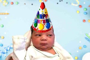Meme of a skeptical baby wearing a colorful birthday hat, surrounded by festive decorations, hilariously questioning the need for yet another birthday celebration.