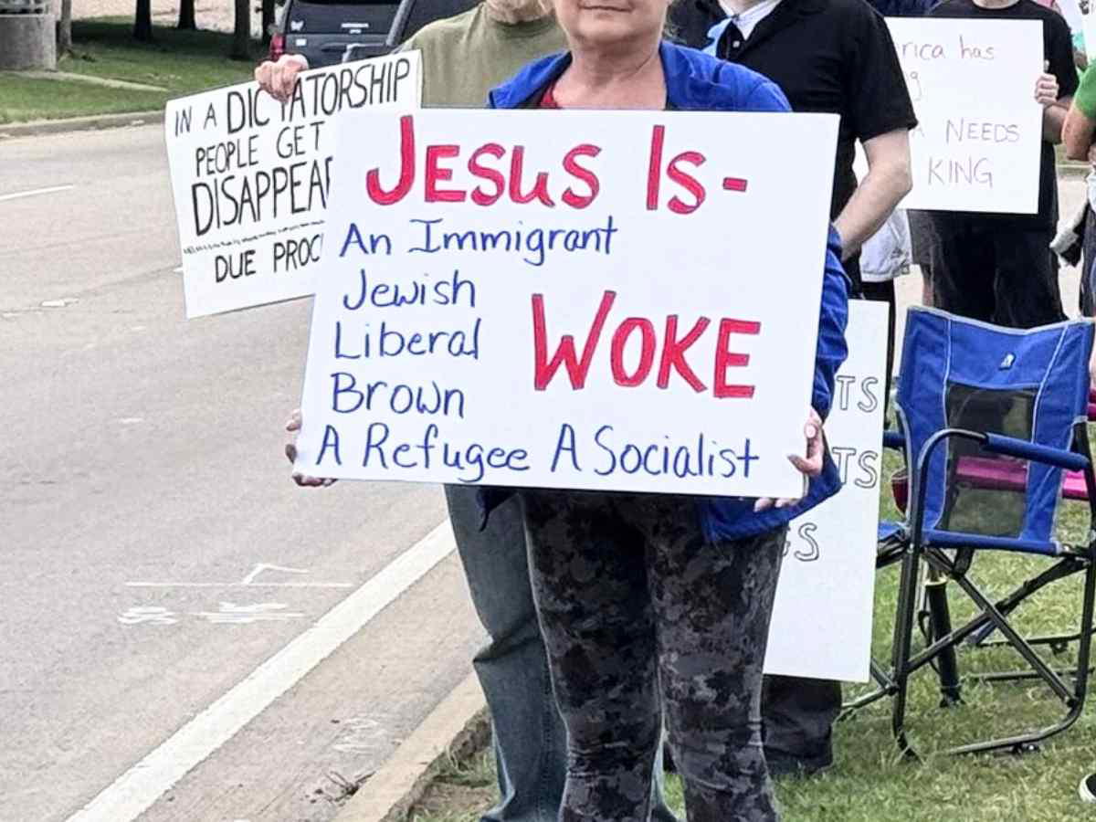 peaceful protest A person holding a protest sign that reads, "JESUS IS - An Immigrant, Jewish, Liberal, Brown, A Refugee, A Socialist," with the word "WOKE" emphasized in red. Other people with signs are visible in the background.